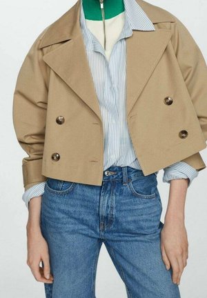 Beige cropped dobbeltspændt jakke over blå stribet skjorte med knapper og grøn top med lynlås, stylet med højtaljede blå jeans.