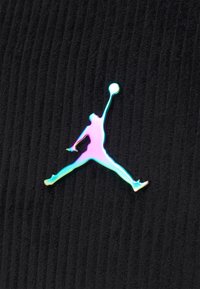 Irydescencyjne logo Jumpman przedstawiające koszykarza sięgającego w górę z piłką na tle czarnej, żebrowanej tkaniny.