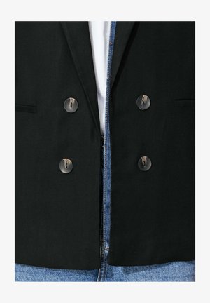 Blazer noir croisé à quatre boutons porté sur une chemise blanche et un jean denim bleu, montrant les détails de la partie médiane.