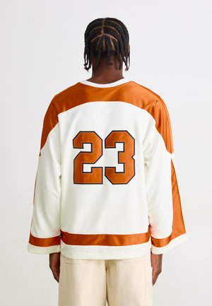 Person with braided hair wearing a white and orange sports jersey with number 23 on the back and light-colored pants, standing facing away.