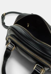 Sac en cuir noir avec une texture lisse, doté de fermetures éclair dorées, d'une ouverture sur le dessus et d'une bandoulière amovible pour le transport. L'intérieur comprend une poche zippée.