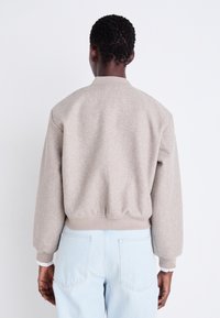 Beez bomber-jakk, millel on pehme tekstuur, soonikugaäär ja -mansetid ning minimalistlik disain. Kombineeritud helesiniste teksadega, näidates tagantvaadet.
