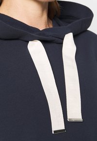Námořnická mikina s texturovanou tkaninou, která má šňůrku s krémovými akcenty a kovovými špičkami. Detail zblízka ukazuje výstřih a šňůrky.