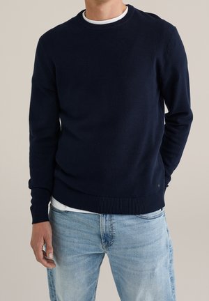 Mann trägt einen dunkelblauen, strukturierten Pullover mit Rundhalsausschnitt über einem weißen Hemd, kombiniert mit hellblauen Denim-Jeans vor einem neutralen Hintergrund.