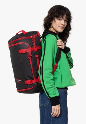 Sac à dos en tissu noir avec des accents rouges, sangles ajustables, fermetures éclair et boucles externes ; porté sur le dos par une personne en pull vert.
