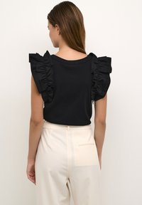 Haut noir à manches volantées et col rond, associé à un pantalon beige clair. Le tissu a une texture lisse.