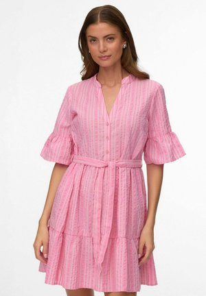 Femme portant une robe rose rayée longueur genou avec des manches courtes volantées et une ceinture nouée à la taille, se tenant devant un fond blanc.