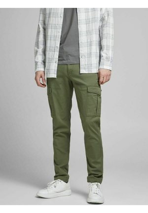 Mężczyzna ubrany w oliwkowozielone spodnie cargo, białe trampki, szarą koszulkę i jasnoszarą otwartą koszulę w kratę stojący na jednolitym tle.