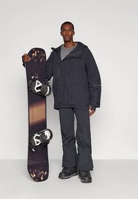Zwarte waterdichte jas met een voorzak en capuchon, gecombineerd met bijpassende broek en zwarte laarzen. Er wordt een snowboard met zwarte bindingen vastgehouden.