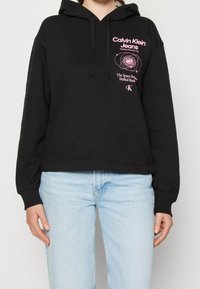 Svart kort hoodie i mjukt tyg, med en rosa grafisk logotyp på bröstet och snörning i huooden. Bärs tillsammans med ljusblå jeans.