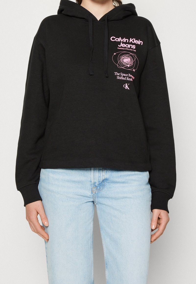 Svart kort hoodie i mjukt tyg, med en rosa grafisk logotyp på bröstet och snörning i huooden. Bärs tillsammans med ljusblå jeans.