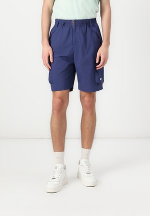Homme portant un short cargo bleu marine, des chaussettes blanches et des baskets blanches, debout devant un fond blanc uni.