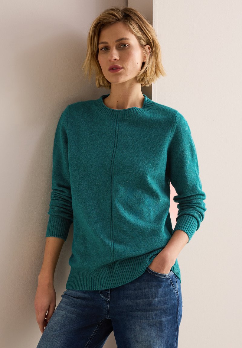 Tyrkis stickad tröja med långa ärmar, rund halsringning, ribbade ärmslut, sidoklämmor och en central sömdetalj. Bärs med blå jeans.