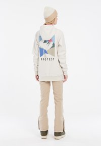 Svijetlosmeđi dukserica s grafičkim dizajnom na leđima koji prikazuje planine i sunce. Uparen s bež hlačama i zelenim čizmama s bočnim patentnim zatvaračem.