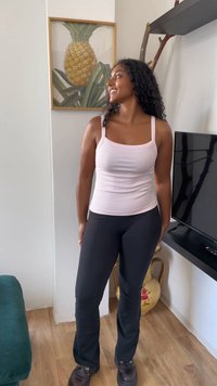 Mulher com cabelo encaracolado, vestindo uma camisola leve rosa e calças pretas, sorri em uma sala com arte de abacaxi e uma televisão.