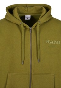Olīvzaļš zip-up kapuce, izgatavota no mīkstas auduma, ar regulējamu kapuci, priekšējām kabatām un izšuvuma logotipu detaļām uz krūtīm.