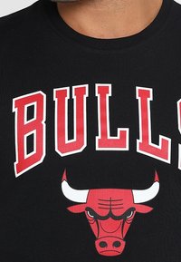 Černé tričko s krátkým rukávem s odvážným bílým nápisem "BULLS" a červenou grafikou hlavy býka s bílými rohy. Hladká bavlněná textura.