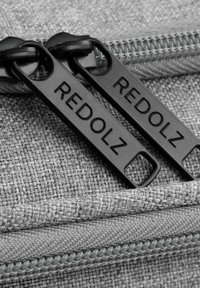 Grijze stoffen tas met zwarte ritstrekkers met het label "REDOLZ." Heeft een gestructureerd materiaal en gestikte naden.