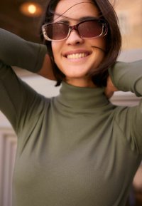Olivinės spalvos raukšlėta polo tipo maikutė ilgomis rankovėmis, derinama su dideliais saulės akiniais, turinčiais skaidrų rėmelį ir tamsius objektyvus.