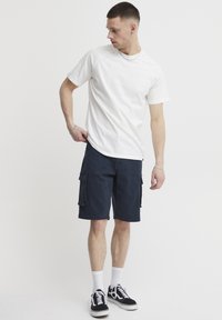 Fehér rövid ujjú póló és tengerészkék cargo rövidnadrág oldalzsebekkel. A modell fekete sportcipőt visel fehér talppal és fehér zoknival.