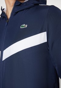 Giacca zip-up navy in tessuto testurizzato, con strisce diagonali bianche e un piccolo logo di coccodrillo verde sul petto.