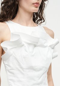 Γυναίκα που φοράει λευκό φόρεμα χωρίς μανίκια με γλυπτικές, κυματιστές λεπτομέρειες υφάσματος κατά μήκος του γραμμής του λαιμού και των ώμων.