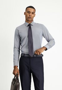Camisa de vestir azul claro con un patrón sutil, combinada con una corbata oscura. Pantalones de traje azul marino a medida y un cinturón trenzado. Sosteniendo un maletín de cuero marrón.