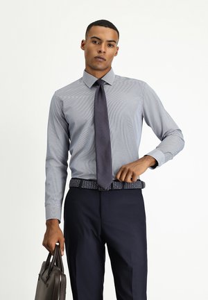 Lichtblauw overhemd met een subtiel patroon, gecombineerd met een donkere stropdas. Op maat gemaakte donkerblauwe broek en een gevlochten riem. Houdt een bruine leren aktetas vast.