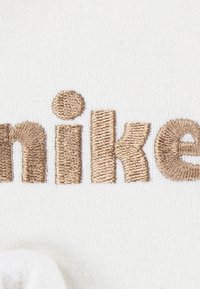 Izšuvums ar vārdu "nike" bēšā diegā uz baltas fona, ar apaļu akcenta punktu virs burta "i."