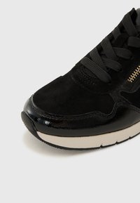 Sneaker noir avec des détails en daim et en cuir laqué, lacets noirs, fermeture éclair sur le côté et semelle en caoutchouc blanc sur fond blanc.