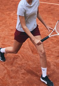 Tennis spiller i bordeaux shorts og grå skjorte, der laver et udfald på grusbanen, mens han holder ketsjeren klar til at ramme bolden.