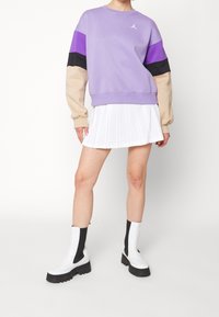 Sudadera de bloques de color púrpura y beige, de corte relajado, con un logo en el pecho, combinada con una falda blanca con plisados y botas de tobillo gruesas en blanco y negro.