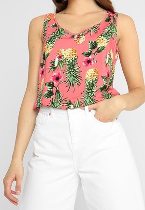 Vrouw draagt een mouwloze roze top met ananas- en bloemenprint, gecombineerd met witte hoge taille broeken, hand in de zak.
