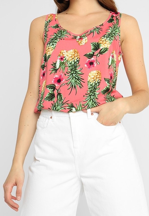Femme portant un haut rose sans manches avec imprimé ananas et fleurs, associé à un pantalon blanc taille haute, main dans la poche.