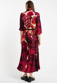 Kobieta w długiej sukience z długim rękawem sięgającej kostek, w odważny abstrakcyjny wzór w kolory czerwonym, czarnym i pomarańczowym, w połączeniu z czarnymi butami na słupku.