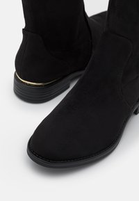 Bottines en daim noir avec un bout arrondi, un talon bas empilé et un subtil accent doré sur le talon. Texture lisse et design épuré.