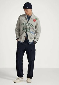 Άντρας που φοράει γκρι καρό σακάκι με εκτύπωση "Yukon Valley", ναυτικές cargo παντελόνες, λευκά αθλητικά παπούτσια, ναυτική σκούφος και τζιν πουκάμισο, στέκεται με τα χέρια στις τσέπες.