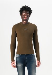 Rippenshirt aus braunem Stoff mit langen Ärmeln, Rundhalsausschnitt und einem kleinen runden Logo auf der Brust, kombiniert mit abgetragenen dunkelblauen Jeans.