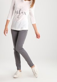 Camisa blanca de manga tres cuartos con texto negro "relax", combinada con pantalones ajustados grises y zapatillas blancas sobre un fondo claro.