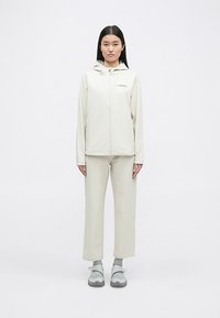 Veste zippée beige clair avec capuche, associée à un pantalon beige assorti. Présente un logo sur la poitrine et une coupe décontractée.