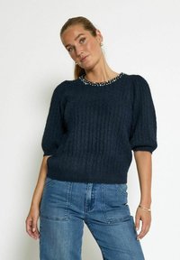 Dark blue knitted sweater with ribbed texture, puffed short sleeves, and a beaded neckline, paired with high-waisted denim jeans.