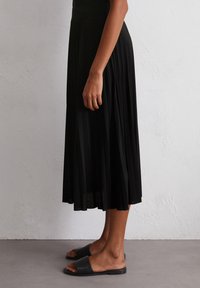 A side view of a person wearing a black pleated skirt and black slide sandals, standing on a neutral-colored floor against a textured wall.