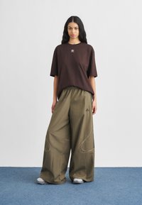 Γυναίκα που στέκεται σε μπλε χαλί, φορώντας μια φαρδιά καφέ κοντομάνικη μπλούζα Adidas και φαρδιά χακί παντελόνια Adidas, με άσπρα αθλητικά παπούτσια.