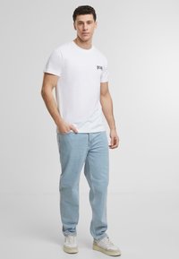 Hvit t-skjorte med korte ermer og "pray" trykt i svart på venstre bryst, sammen med lyseblå jeans og beige sneakers.