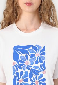 T-shirt blanc avec un design graphique floral bleu et des accents roses. Encolure ronde et manches courtes ; texture de tissu lisse.