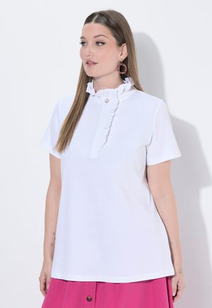 Vrouw met lang, stijl haar die een witte blouse met geplooide hals en korte mouwen draagt en een roze rok, kijkend naar links met zichtbare oorbellen.