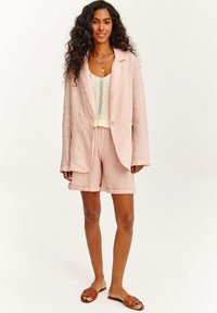 Světle růžový lněný blejzr přes krémový pletený top, doplněný o odpovídající lněné šortky a hnědé skládací sandály. Model stojí na neutrálním pozadí.