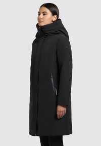 Naine kannab pikka musta talvekatet, millel on kõrge krae ja kapuuts, seistes külg ees lihtsa heleda tausta ees.