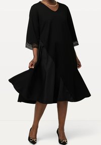 Černé šaty s výstřihem do V, tříčtvrtečními rukávy a čipkovým lemováním. Silueta ve tvaru písmene A s volánovou sukni. Nosené s černými podpatky se zlatými akcenty.