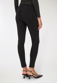 Donna che indossa jeans skinny neri e tacchi slingback neri, vista da dietro su uno sfondo bianco semplice.